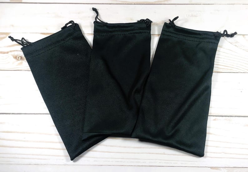 May include: Three black drawstring pouches are arranged on a light wood-grain surface. Each pouch is made of a smooth, dark fabric and has a drawstring closure at the top. The pouches are empty and ready for use.