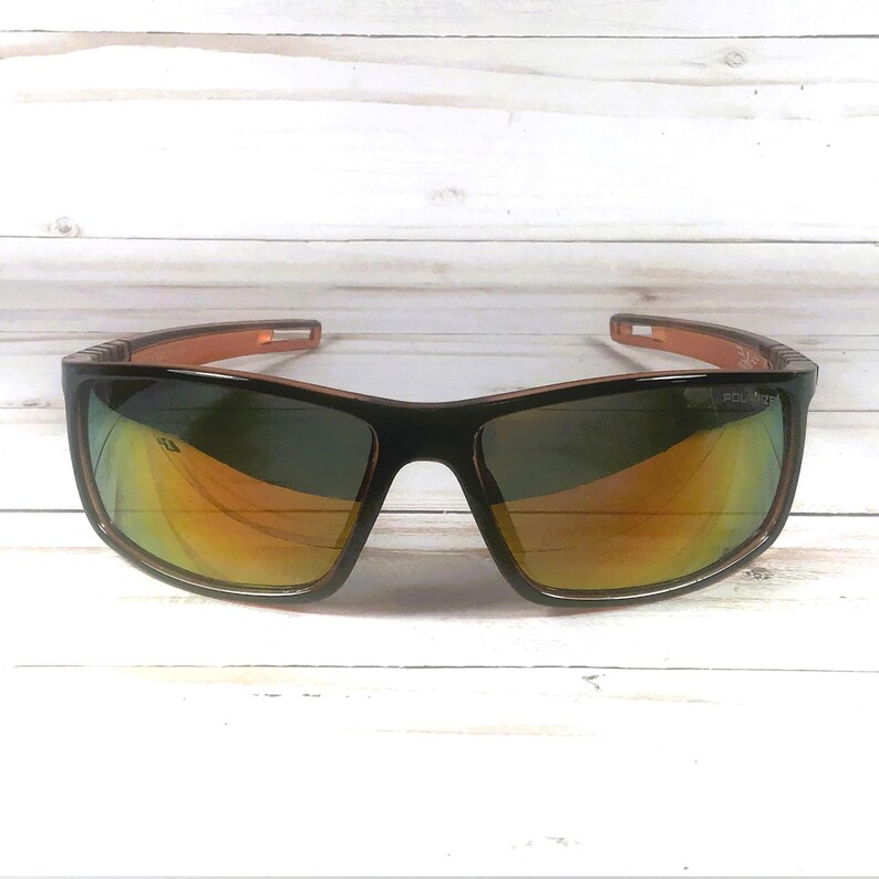 May include: Black sunglasses with orange and green reflective lenses. The frames are black with orange accents on the arms. The lenses have a polarized coating. The sunglasses are set against a white wooden background.