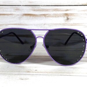 Peut inclure: Lunettes de soleil aviateur violettes avec des verres fonc&eacute;s. Les montures sont d'un violet vif, et de petits strass argent&eacute;s sont sertis le long des bords ext&eacute;rieurs des verres. Les lunettes ont un design classique &agrave; double pont.