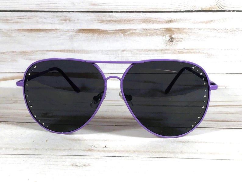 Peut inclure: Lunettes de soleil aviateur violettes avec des verres fonc&eacute;s. Les montures sont d'un violet vif, et de petits strass argent&eacute;s sont sertis le long des bords ext&eacute;rieurs des verres. Les lunettes ont un design classique &agrave; double pont.