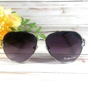 May include: Silver-framed aviator sunglasses with dark purple lenses. The sunglasses have a double bridge and the text "Ruby Lee" is printed on the right lens. Yellow flowers and green leaves are in the background.