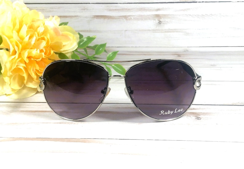 May include: Silver-framed aviator sunglasses with dark purple lenses. The sunglasses have a double bridge and the text "Ruby Lee" is printed on the right lens. Yellow flowers and green leaves are in the background.