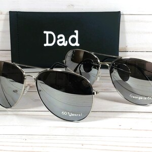 Może przedstawiać: Dwie pary srebrnych okular&oacute;w przeciwsłonecznych typu aviator z lustrzanymi szkłami. Jedna para ma napis "Dad" na etui, a druga para ma napis "60 Years!" na jednym szkle i "George is 60" na drugim szkle.