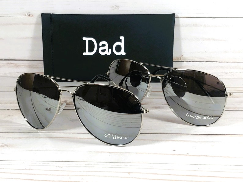 Może przedstawiać: Dwie pary srebrnych okular&oacute;w przeciwsłonecznych typu aviator z lustrzanymi szkłami. Jedna para ma napis "Dad" na etui, a druga para ma napis "60 Years!" na jednym szkle i "George is 60" na drugim szkle.