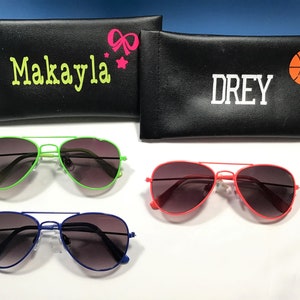 May include: Three pairs of aviator sunglasses with different colored frames: green, red, and blue. Each pair has dark tinted lenses. The sunglasses are displayed on a white surface with two black zippered cases behind them. One case has the name "Makayla" and a pink bow and star. The other case has the name "Drey" and a basketball.
