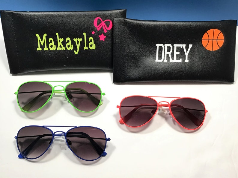 May include: Three pairs of aviator sunglasses with different colored frames: green, red, and blue. Each pair has dark tinted lenses. The sunglasses are displayed on a white surface with two black zippered cases behind them. One case has the name "Makayla" and a pink bow and star. The other case has the name "Drey" and a basketball.