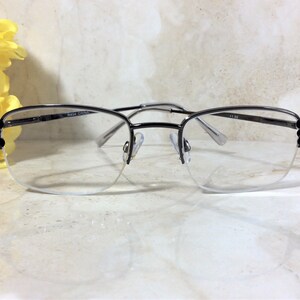 May include: Semi-rimless eyeglasses with a silver metal frame. The lenses are clear, and the frame has decorative black accents on the temples. The glasses are set against a marble-like surface with yellow flowers in the background.
