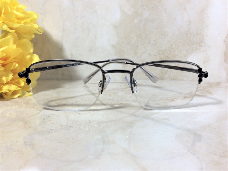 May include: Semi-rimless eyeglasses with a silver metal frame. The lenses are clear, and the frame has decorative black accents on the temples. The glasses are set against a marble-like surface with yellow flowers in the background.