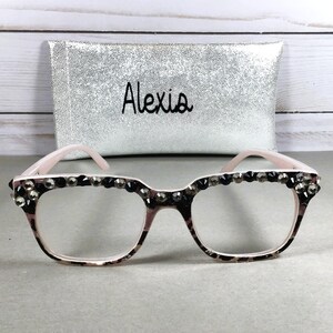 May include: Pink-framed eyeglasses with a leopard print pattern and embellished with dark rhinestones along the top. A silver clutch bag with the name "Alexia" is in the background.