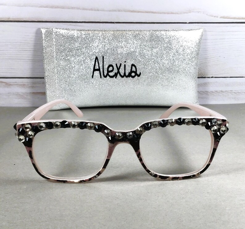 May include: Pink-framed eyeglasses with a leopard print pattern and embellished with dark rhinestones along the top. A silver clutch bag with the name "Alexia" is in the background.
