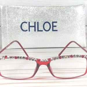 May include: Red-framed eyeglasses with decorative rhinestones along the top rim. The glasses are in front of a silver, textured case with the name "CHLOE" in navy blue lettering.