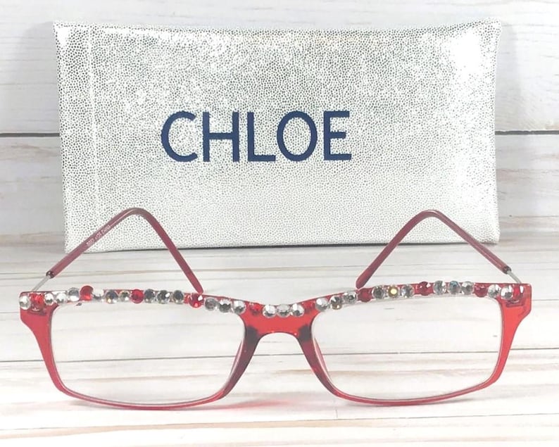 May include: Red-framed eyeglasses with decorative rhinestones along the top rim. The glasses are in front of a silver, textured case with the name "CHLOE" in navy blue lettering.