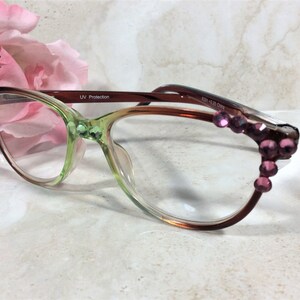 May include: A pair of eyeglasses with a gradient green and brown frame. The frames are adorned with pink gemstones. The lenses are clear, and the glasses have a UV protection label. The glasses are set against a soft background with a pink flower.