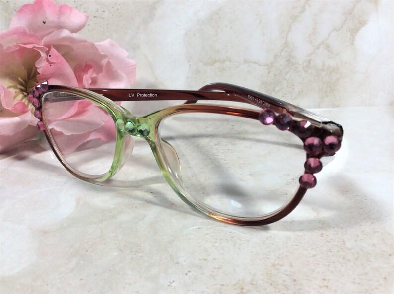 May include: A pair of eyeglasses with a gradient green and brown frame. The frames are adorned with pink gemstones. The lenses are clear, and the glasses have a UV protection label. The glasses are set against a soft background with a pink flower.