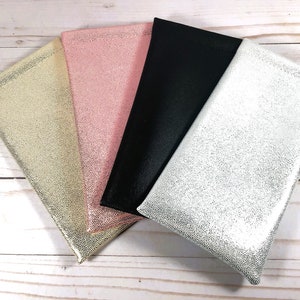 May include: Four different colored fabric pouches with a textured surface. The pouches are arranged in a stack with the silver pouch on the bottom, followed by the black pouch, the pink pouch, and the gold pouch on top.