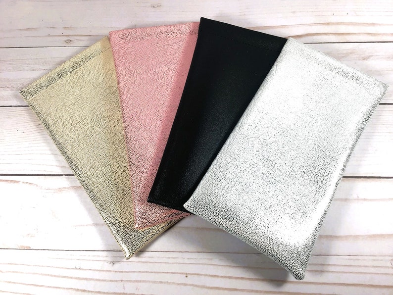 May include: Four different colored fabric pouches with a textured surface. The pouches are arranged in a stack with the silver pouch on the bottom, followed by the black pouch, the pink pouch, and the gold pouch on top.