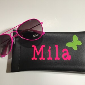 May include: Pink aviator sunglasses with a black case that has the name "Mila" and a green butterfly printed on it.