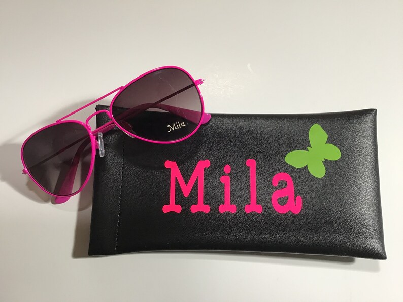 May include: Pink aviator sunglasses with a black case that has the name "Mila" and a green butterfly printed on it.