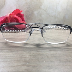 May include: Black and silver-toned eyeglasses with a half-rim design. The glasses have a rectangular shape and clear lenses. The frame is black on top and silver on the bottom. A red rose and decorative beads are in the background.