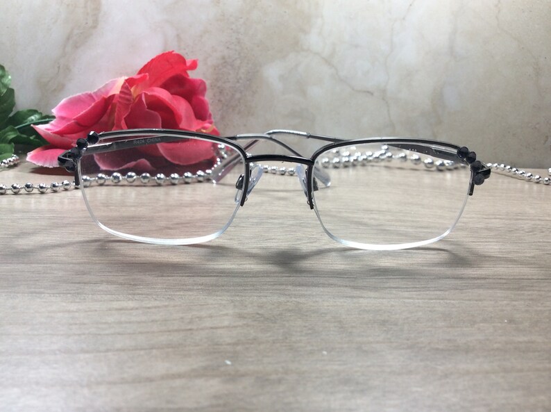 May include: Black and silver-toned eyeglasses with a half-rim design. The glasses have a rectangular shape and clear lenses. The frame is black on top and silver on the bottom. A red rose and decorative beads are in the background.