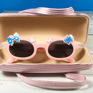 May include: Pink sunglasses with dark lenses, adorned with blue flower and butterfly details. The sunglasses are presented in a matching pink striped case with a handle. The case is open, displaying the sunglasses.