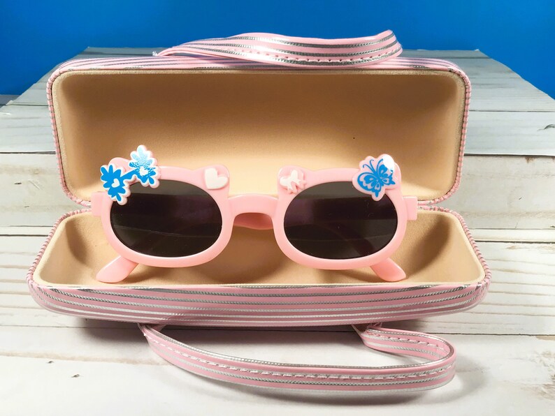 May include: Pink sunglasses with dark lenses, adorned with blue flower and butterfly details. The sunglasses are presented in a matching pink striped case with a handle. The case is open, displaying the sunglasses.