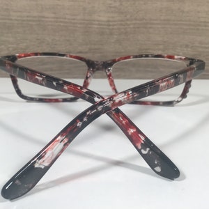 May include: Eyeglasses with a marbled pattern in red, black, and white. The frames have a rectangular shape and clear lenses. The temples curve back, and the glasses are set against a white surface.