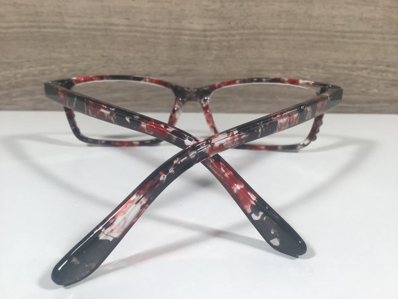 May include: Eyeglasses with a marbled pattern in red, black, and white. The frames have a rectangular shape and clear lenses. The temples curve back, and the glasses are set against a white surface.