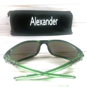 May include: Green sunglasses with dark lenses and a black case. The case is black with the name "Alexander" in white lettering. The sunglasses have a modern design.