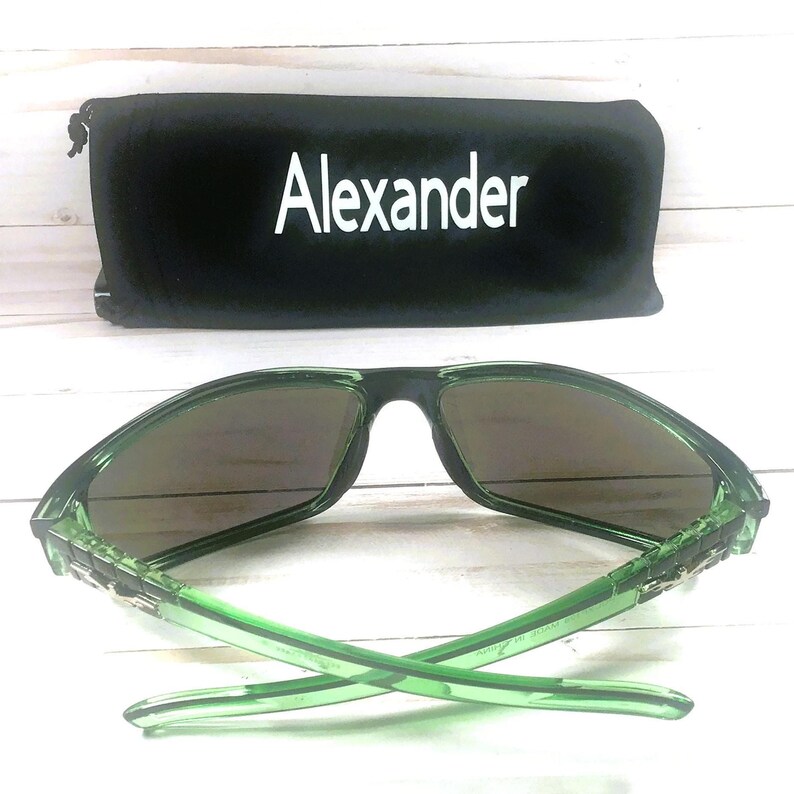 May include: Green sunglasses with dark lenses and a black case. The case is black with the name "Alexander" in white lettering. The sunglasses have a modern design.