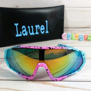 Puede incluir: Un par de gafas de sol rosa y azul con pintura negra salpicada. Las gafas tienen una lente espejada arco&iacute;ris y est&aacute;n sobre una superficie de madera blanca. La palabra "Laurel" est&aacute; escrita en una caja negra en el fondo.