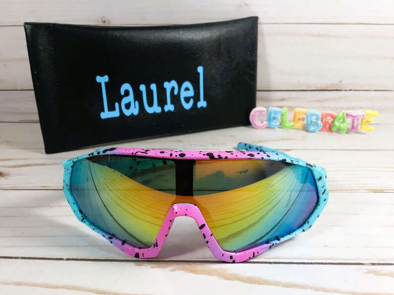 Puede incluir: Un par de gafas de sol rosa y azul con pintura negra salpicada. Las gafas tienen una lente espejada arco&iacute;ris y est&aacute;n sobre una superficie de madera blanca. La palabra "Laurel" est&aacute; escrita en una caja negra en el fondo.