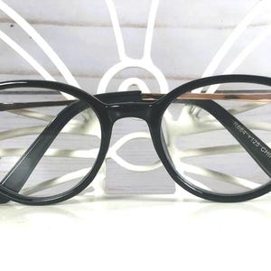 May include: Black oval-shaped spectacles featuring clear lenses and thin metallic temples. The outer corners of the black frames are embellished with three small, clear, circular rhinestones on each side. The inner temple arm displays the text R884 FT25 CHINA.