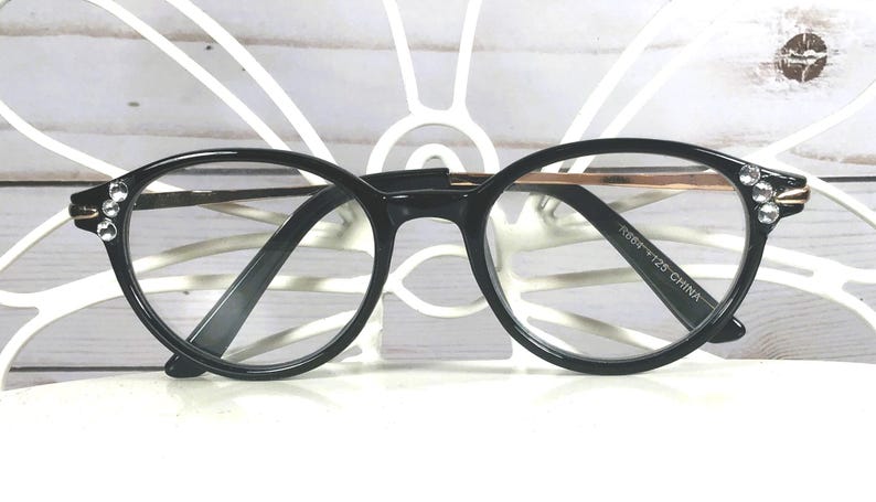 May include: Black oval-shaped spectacles featuring clear lenses and thin metallic temples. The outer corners of the black frames are embellished with three small, clear, circular rhinestones on each side. The inner temple arm displays the text R884 FT25 CHINA.