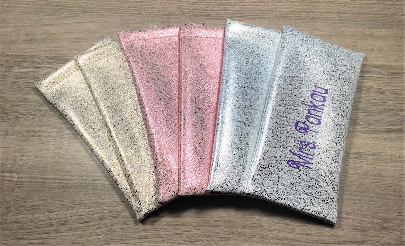 Pu&ograve; includere: Una collezione di pochette metallizzate nei toni dell'oro, del rosa e dell'argento, disposte su una superficie di legno. Una pochette &egrave; ricamata con il nome "Mrs Pankau" in viola.