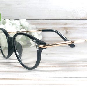 May include: Black round-frame glasses featuring thin gold-tone metal temples and clear lenses. The outer corners of the black plastic frames are accented with two small, clear, sparkling rhinestones on each side, adding a subtle decorative detail.