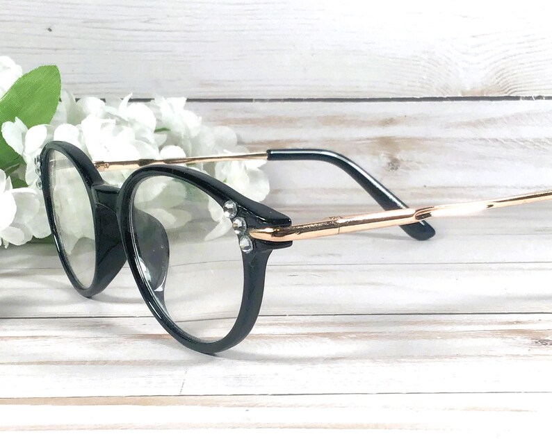 May include: Black round-frame glasses featuring thin gold-tone metal temples and clear lenses. The outer corners of the black plastic frames are accented with two small, clear, sparkling rhinestones on each side, adding a subtle decorative detail.