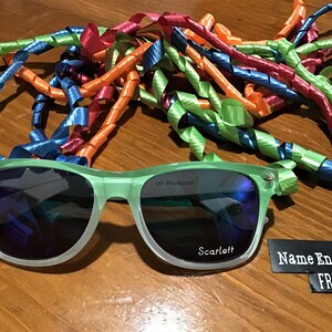 May include: Green sunglasses with UV protection and the name "Scarlett" engraved on the right lens. A black and white label reads "Name Engraved FREE".