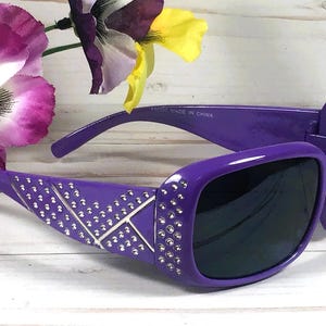 May include: Purple sunglasses with a square frame and dark lenses. The arms are decorated with silver-colored rhinestones in a geometric pattern. The sunglasses are set against a background with purple and yellow flowers.