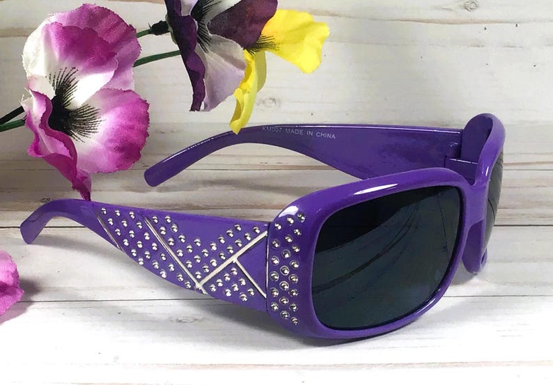 May include: Purple sunglasses with a square frame and dark lenses. The arms are decorated with silver-colored rhinestones in a geometric pattern. The sunglasses are set against a background with purple and yellow flowers.