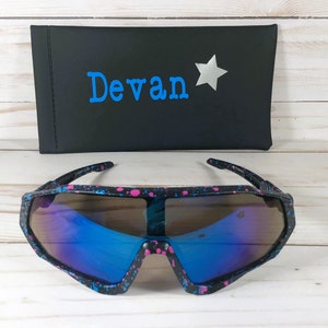 May include: Black sunglasses with a pink and blue paint splatter pattern and reflective blue lenses. Behind the sunglasses is a black rectangular case featuring the name Devan in blue text next to a silver star graphic.