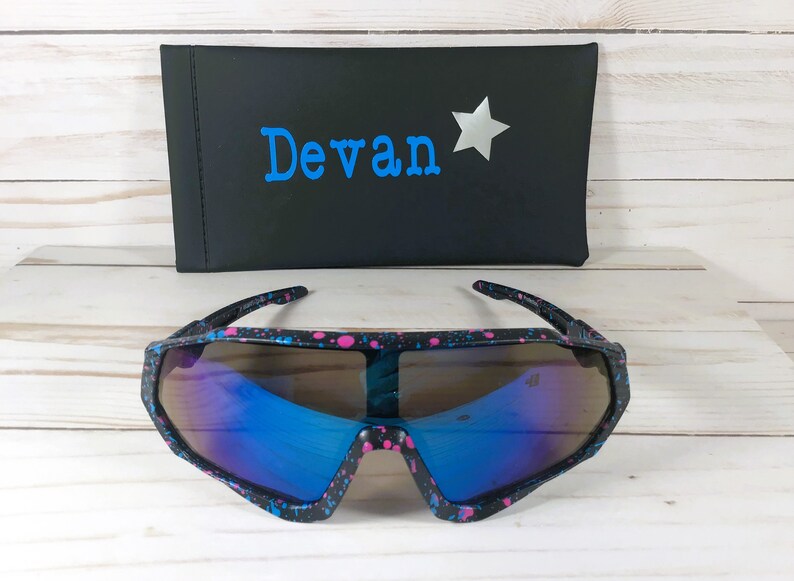 May include: Black sunglasses with a pink and blue paint splatter pattern and reflective blue lenses. Behind the sunglasses is a black rectangular case featuring the name Devan in blue text next to a silver star graphic.
