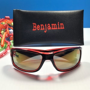 May include: Red sunglasses with a black frame and mirrored lenses. The sunglasses are in a black case with the name "Benjamin" printed in red.