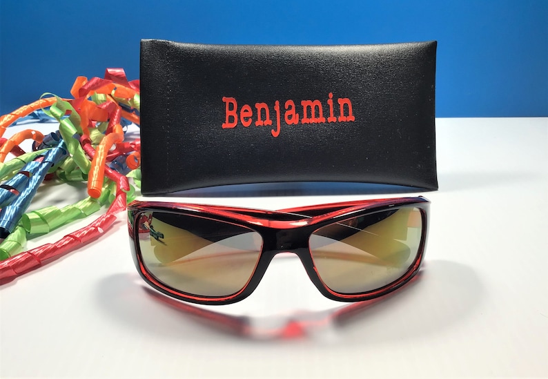 May include: Red sunglasses with a black frame and mirrored lenses. The sunglasses are in a black case with the name "Benjamin" printed in red.