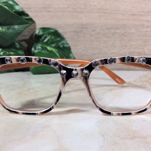 Puede incluir: Gafas de montura cuadrada con estampado de leopardo, adornadas con pedrer&iacute;a transparente en el borde superior. El interior y las patillas son de color marr&oacute;n claro. Las lentes son transparentes.