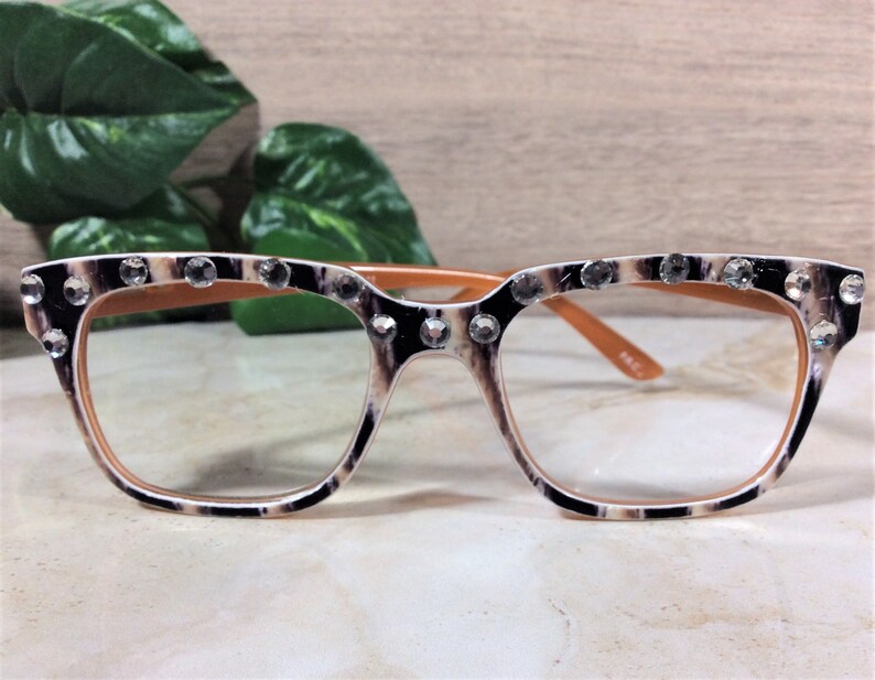 Puede incluir: Gafas de montura cuadrada con estampado de leopardo, adornadas con pedrer&iacute;a transparente en el borde superior. El interior y las patillas son de color marr&oacute;n claro. Las lentes son transparentes.