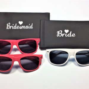 May include: Two pairs of sunglasses with black lenses. One pair is red and the other is white. The sunglasses come with black cases that say "Bride" and "Bridesmaid" in white text.