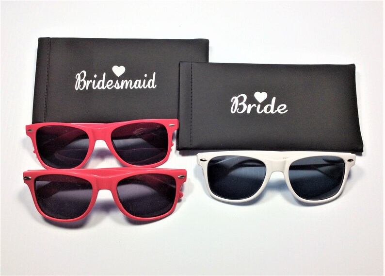 May include: Two pairs of sunglasses with black lenses. One pair is red and the other is white. The sunglasses come with black cases that say "Bride" and "Bridesmaid" in white text.
