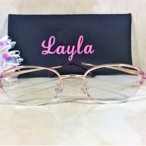 Pu&ograve; includere: Occhiali color oro rosa con lenti trasparenti e dettagli di pietre preziose viola sulle aste. Gli occhiali sono davanti a un astuccio nero con il nome "Layla" in corsivo rosa.