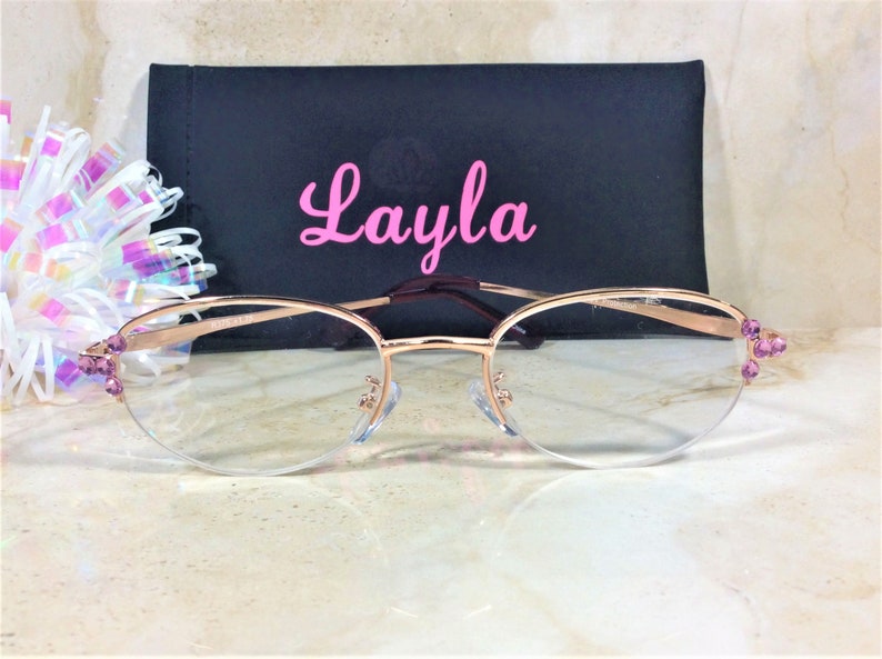 Pu&ograve; includere: Occhiali color oro rosa con lenti trasparenti e dettagli di pietre preziose viola sulle aste. Gli occhiali sono davanti a un astuccio nero con il nome "Layla" in corsivo rosa.